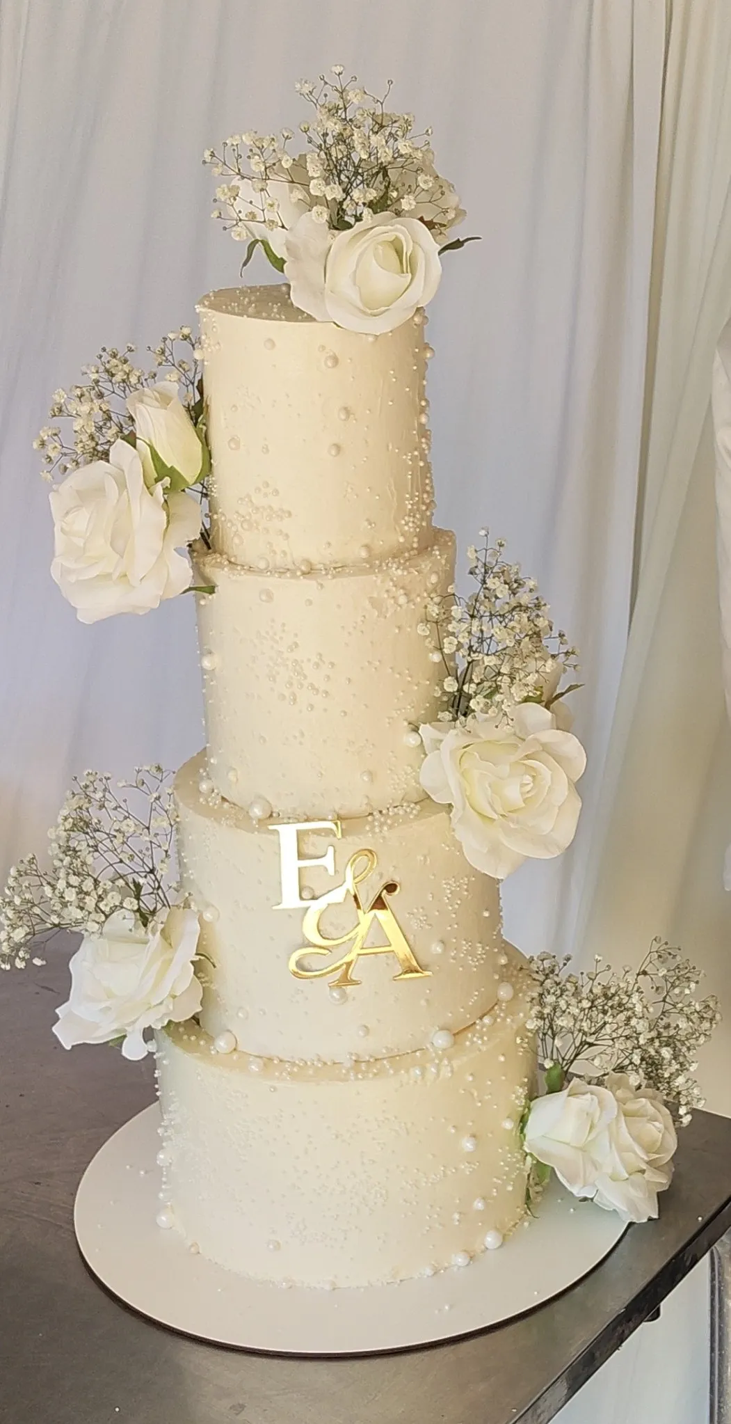 Tarta blanca de cuatro pisos con perlas, rosas blancas e iniciales doradas